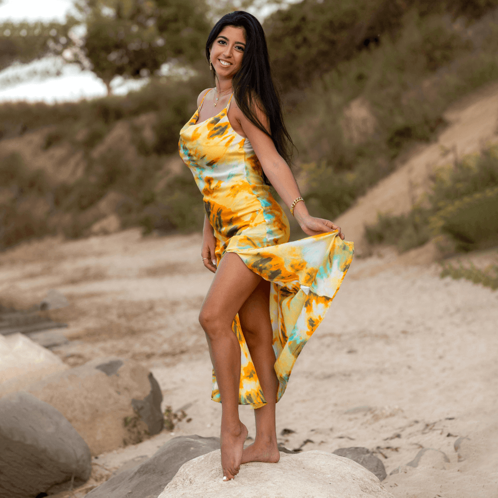 Model wearing a vibrant yellow and blue tie-dye dress on a beach, enjoying the summer day.